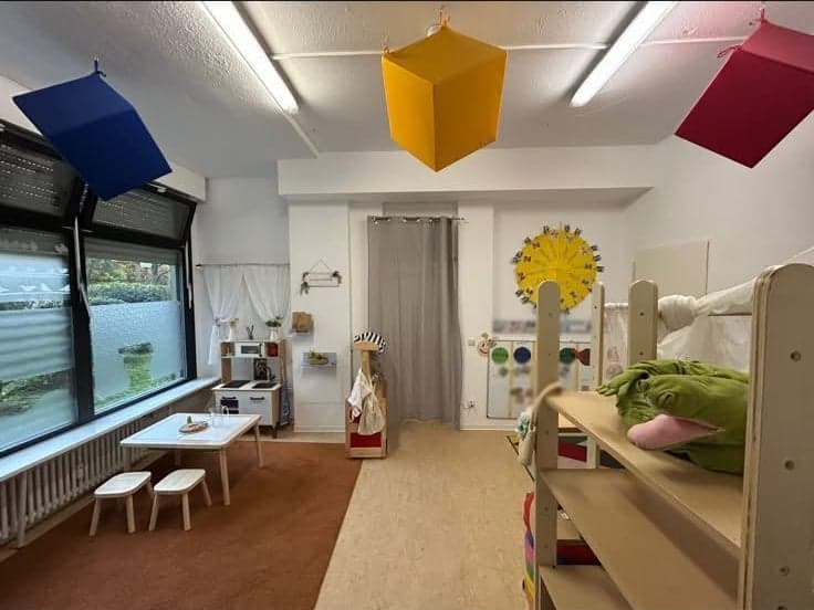 Gruppenraum mit bunten Würfeln unter der Decke, einem kleinen Tisch mit Hockern, einer Spielküche und einem Hochbett aus Holz