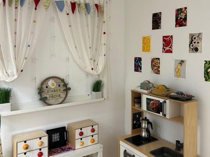 Spielküche aus Holz unter Fotos von Obst, daneben ein Fenster mit Wimpelvorhang und dem Schild „Kinderküche“