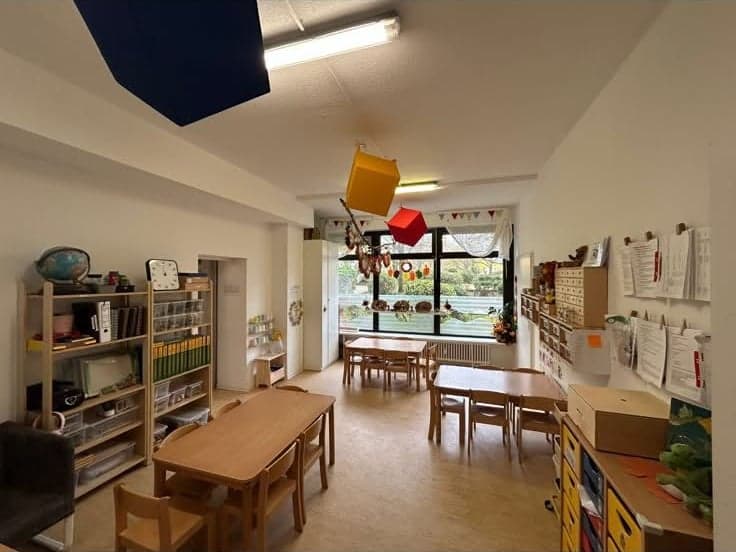 Langer Raum mit Kindertischen, Holzregalen und einem großen Fenster ins Grüne