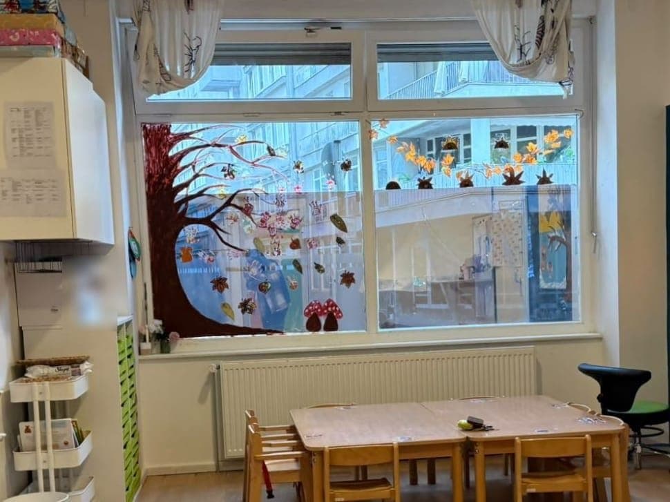 Gruppenraum mit Kindertischen vor einem großen, herbstlich bemalten Fenster
