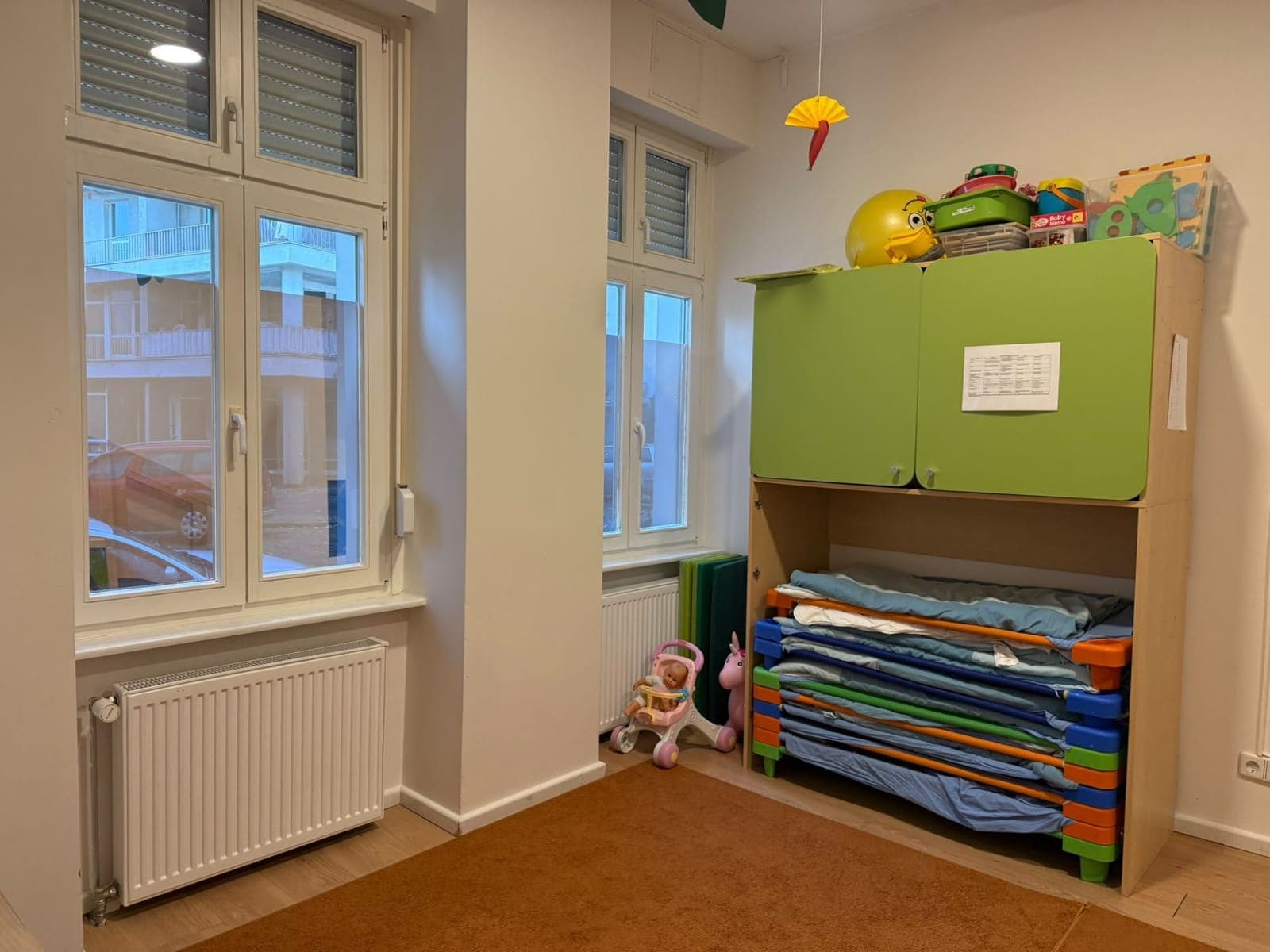 Gestapelte Kinderliegen mit Decken unter einem grünen Schrank, daneben hohe Fenster